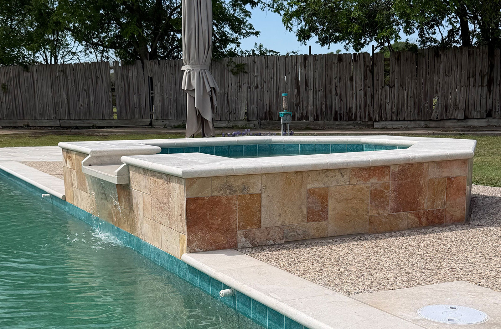A raised stone hot tub with a waterfall feature overflows into a swimming pool, set in a backyard with a wooden fence and green trees.