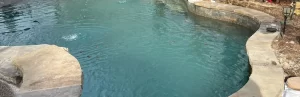 Irregularly shaped backyard swimming pool with stone coping, surrounded by rocks and some outdoor items visible at the poolside.