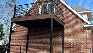 A brick house features a second-story balcony with black metal railings and supports, extending outward from the upper floor.