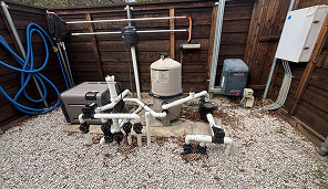 Outdoor pool equipment area with various pipes, valves, a filter tank, heater, and electrical boxes on gravel, enclosed by a wooden fence.