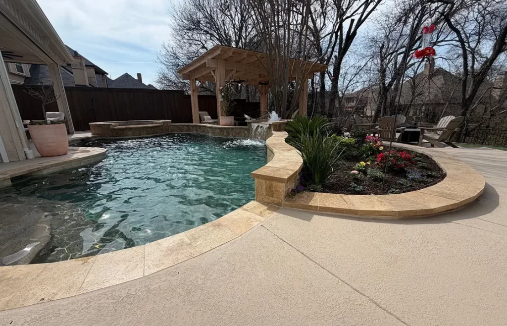 Backyard with a swimming pool, stone edging, adjacent flower bed, pergola seating area, waterfall feature, and trees in the background.