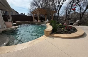 Backyard with a swimming pool, stone edging, adjacent flower bed, pergola seating area, waterfall feature, and trees in the background.