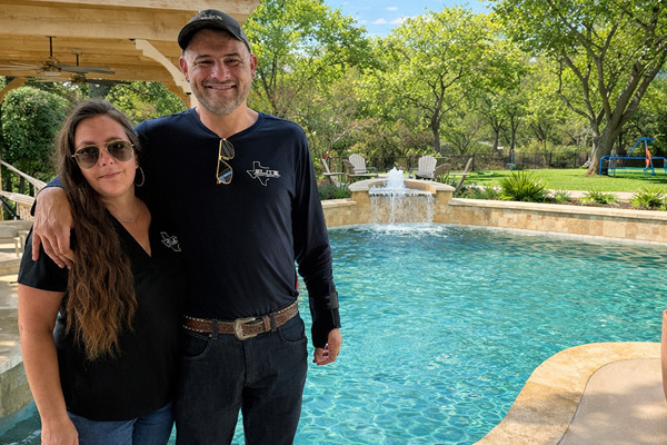 A man and woman stand by an outdoor swimming pool, with trees, lawn chairs, and a fountain visible in the background on a sunny day.