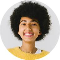 A person with short curly hair, wearing a yellow sweater and white shirt, smiling at the camera against a plain light background.