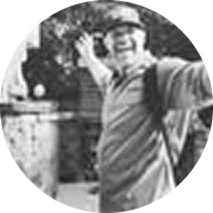 A person wearing a hat and backpack smiles with arms outstretched while standing outdoors. The image is in black and white.