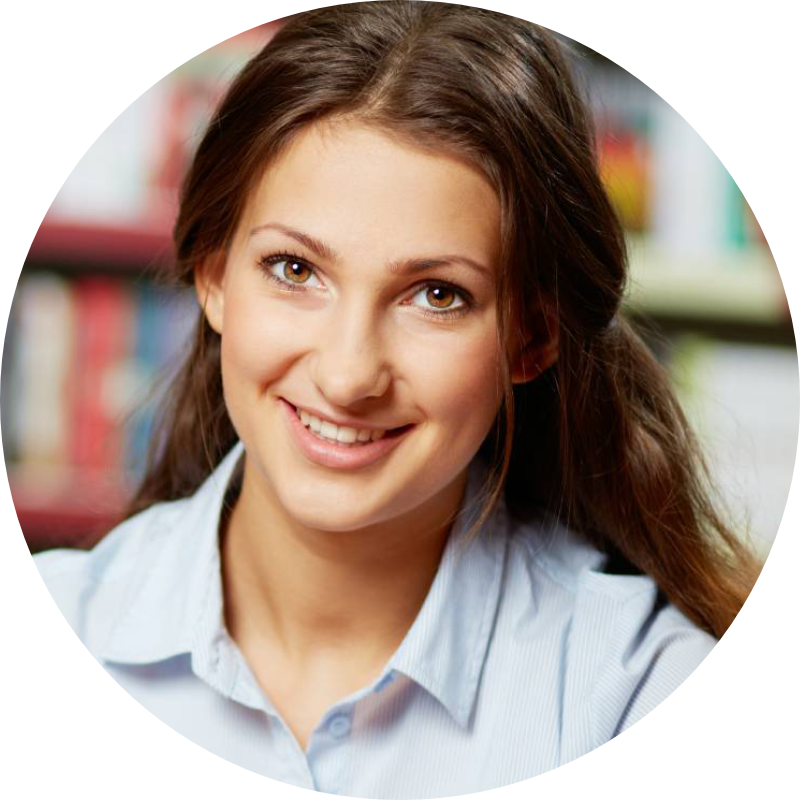 A young woman with long brown hair, wearing a light blue shirt, smiling in front of a blurred background of bookshelves.