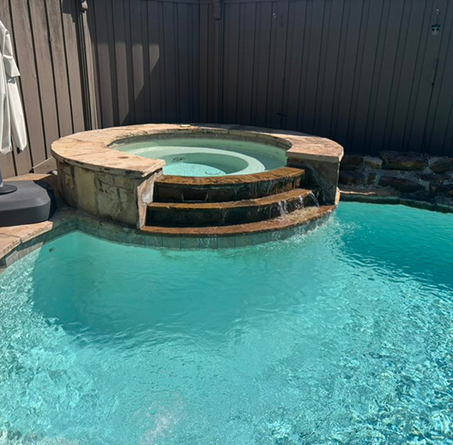 A round hot tub with water flowing into a clear blue swimming pool, surrounded by a brown wooden fence and stone edging.