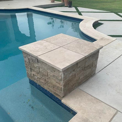 A stone block extends from the pool deck into the swimming pool, partially surrounded by water, with grass and potted plants visible in the background.