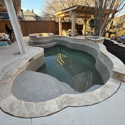 Partially filled backyard pool with attached hot tub, surrounded by stone tiles, near a covered patio and wooden fence. Two people stand in the background.