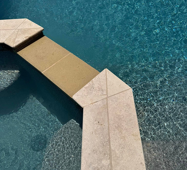 A stone walkway crosses over a clear blue swimming pool, connecting two sections above the water. Sunlight creates patterns on the pool's surface.