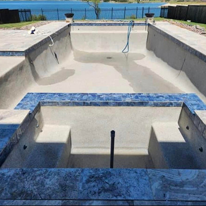 An empty in-ground swimming pool with some water at the bottom, steps leading in, and a view of a lake and fence in the background.