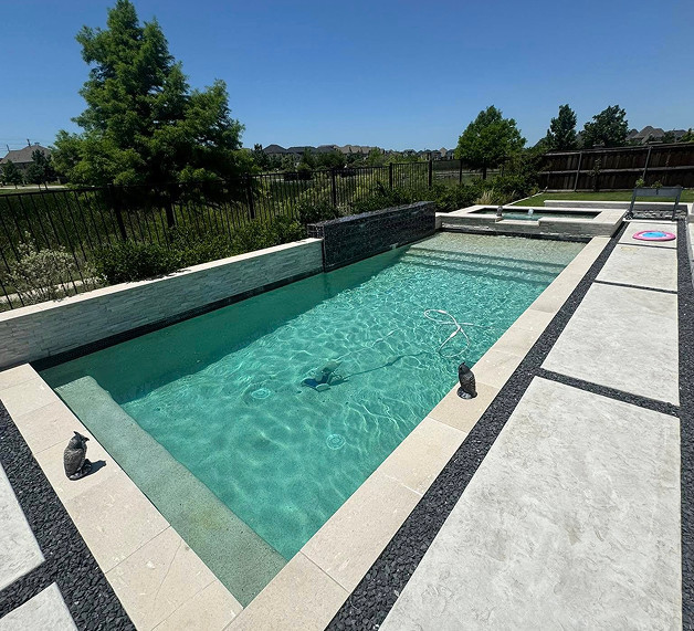 Rectangular backyard swimming pool with clear water, surrounded by light stone tiles, two small statues on the edge, and a fenced yard with trees and grass in the background.
