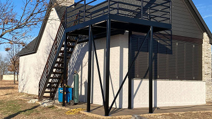 A black metal staircase and landing structure is attached to the exterior of a light brick building near a tree, with equipment and a yellow hose below.