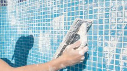 A person applies grout to blue mosaic tiles using a float tool.
