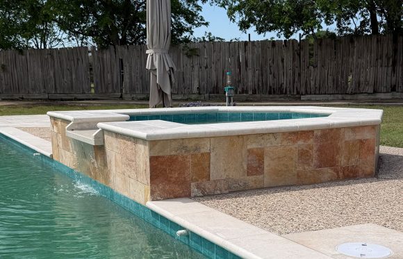 A raised stone hot tub with a waterfall feature overflows into a swimming pool, set in a backyard with a wooden fence and green trees.