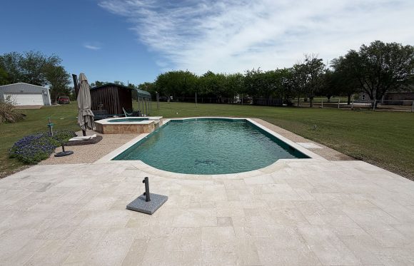 A rectangular outdoor swimming pool with an attached hot tub, surrounded by a stone patio, in a spacious backyard with grass, trees, and outbuildings under a partly cloudy sky.
