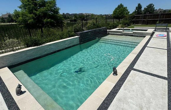 Rectangular backyard swimming pool with clear water, surrounded by light stone tiles, two small statues on the edge, and a fenced yard with trees and grass in the background.