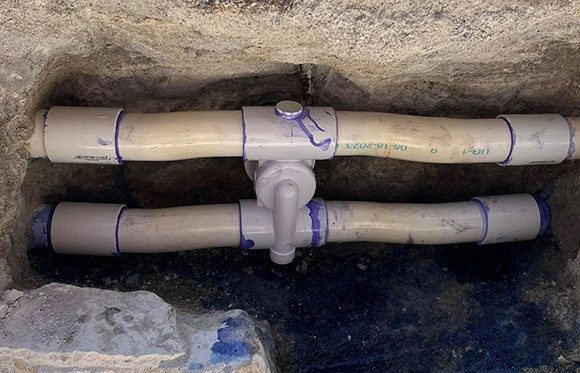 Two parallel PVC pipes connected by a central valve, set in a cutout section of concrete or stone.