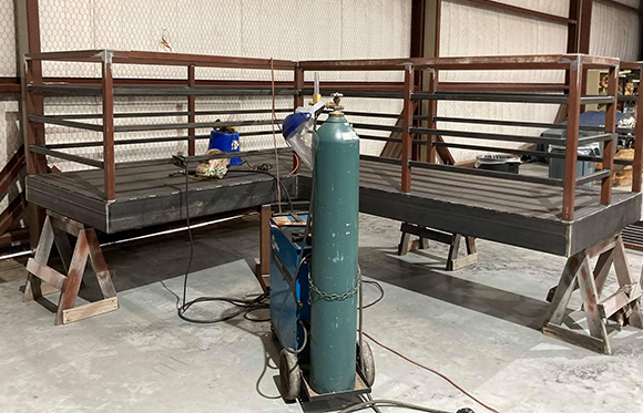 Two metal platforms with railings being welded in a workshop, with welding equipment and gas cylinder positioned in front.
