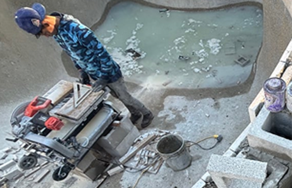 A worker in a blue camouflage hoodie operates a tile saw next to an unfinished pool partially filled with murky water and surrounded by construction materials.