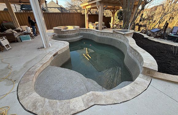 Unfinished backyard pool with a small amount of water, surrounded by stone coping, patio area, and nearby pergola; construction materials are visible.