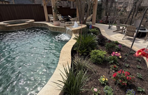 A backyard pool with a stone edge, small waterfall, adjacent flower bed, pergola with seating area, and trees in the background—perfect for enjoying outdoor living spaces in North Texas.