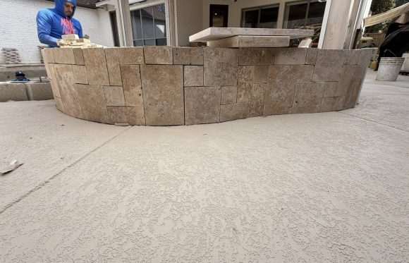 A person in a blue hoodie works on constructing a curved stone wall on a textured patio, enhancing outdoor living spaces North Texas under a covered area.
