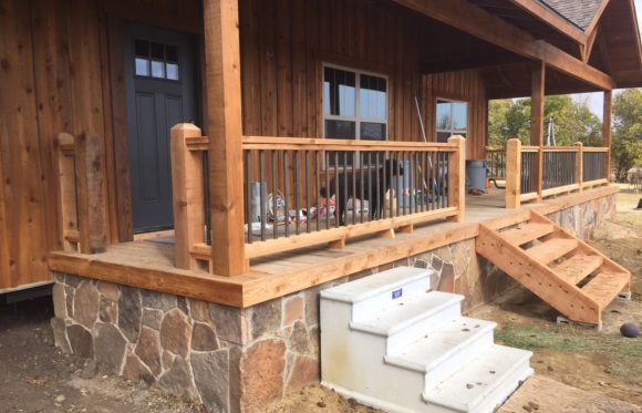 A wooden porch with a stone base, black spindle railing, wooden posts, steps on both sides, and various items scattered on the deck—perfect inspiration for home renovations in North Texas.