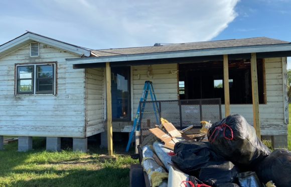 A small, weathered house in North Texas under renovation with a blue ladder, exposed beams, and construction debris—including black trash bags—piled in the yard showcases the realities of home renovations.